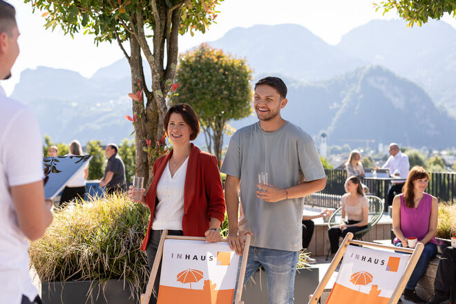 Ein Mann und eine Frau stehen lächelnd neben Liegestühlen und unterhalten sich mit einer weiteren Person. Im Hintergrund genießen andere Menschen die sonnige Atmosphäre mit Bergblick. Ein Mann und eine Frau stehen lächelnd neben Liegestühlen und unterhalten sich mit einer weiteren Person. Im Hintergrund genießen andere Menschen die sonnige Atmosphäre mit Bergblick.
