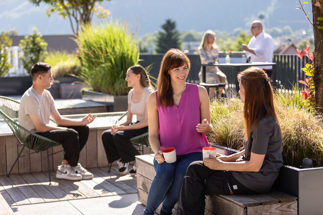 Menschen sitzen draußen und plaudern entspannt mit Getränken in der Hand. Die Umgebung ist grün und sonnig, mit einer malerischen Berglandschaft im Hintergrund. Menschen sitzen draußen und plaudern entspannt mit Getränken in der Hand. Die Umgebung ist grün und sonnig, mit einer malerischen Berglandschaft im Hintergrund.