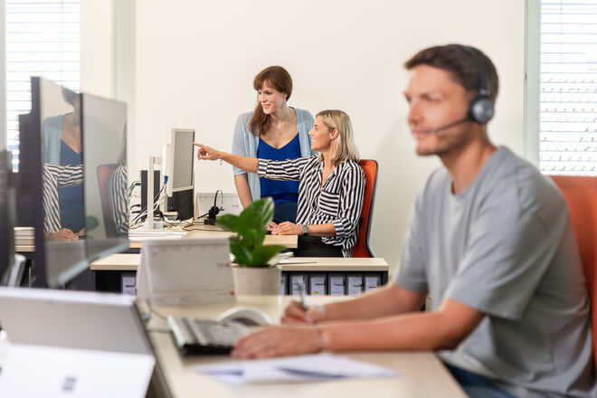 Zwei Frauen besprechen Inhalte am Bildschirm, während ein Mann mit Headset konzentriert am Schreibtisch arbeitet. Zwei Frauen besprechen Inhalte am Bildschirm, während ein Mann mit Headset konzentriert am Schreibtisch arbeitet.