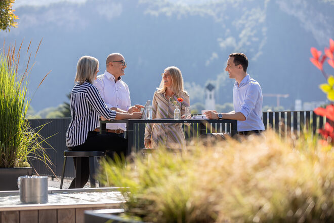 Vier Personen sitzen an einem Tisch im Freien und unterhalten sich. Die Szenerie ist sonnig, mit einer Berglandschaft und Blumen im Hintergrund. Vier Personen sitzen an einem Tisch im Freien und unterhalten sich. Die Szenerie ist sonnig, mit einer Berglandschaft und Blumen im Hintergrund.