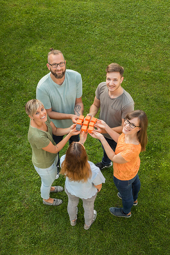Fünf Personen stehen im Kreis auf einer Wiese, halten orange Würfel in der Mitte und lächeln nach oben, als ob sie an einem Teamspiel teilnehmen. Fünf Personen stehen im Kreis auf einer Wiese, halten orange Würfel in der Mitte und lächeln nach oben, als ob sie an einem Teamspiel teilnehmen.