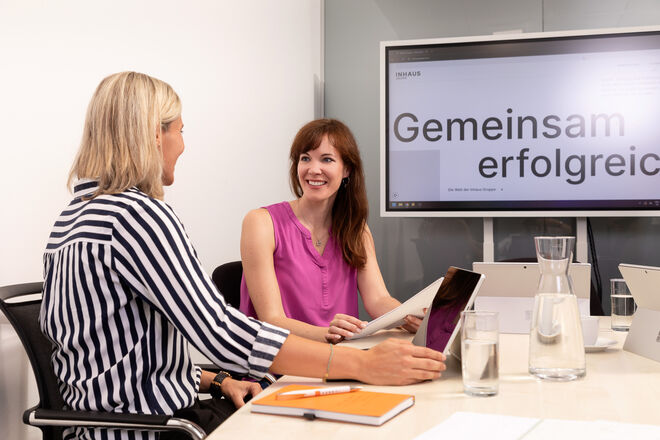 Zwei Frauen lächeln und sprechen in einem modernen Büro, umgeben von Laptops und Notizen, mit einem Bildschirm im Hintergrund. Zwei Frauen lächeln und sprechen in einem modernen Büro, umgeben von Laptops und Notizen, mit einem Bildschirm im Hintergrund.