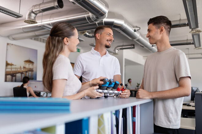 Drei Personen stehen an einem Tresen und unterhalten sich freundlich, während sie technische Bauteile betrachten und diskutieren Drei Personen stehen an einem Tresen und unterhalten sich freundlich, während sie technische Bauteile betrachten und diskutieren