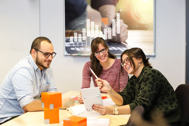 Drei Personen sitzen an einem Tisch und diskutieren fröhlich, mit Dokumenten und einem Tablet. Im Hintergrund ist ein Diagrammposter zu sehen. Drei Personen sitzen an einem Tisch und diskutieren fröhlich, mit Dokumenten und einem Tablet. Im Hintergrund ist ein Diagrammposter zu sehen.